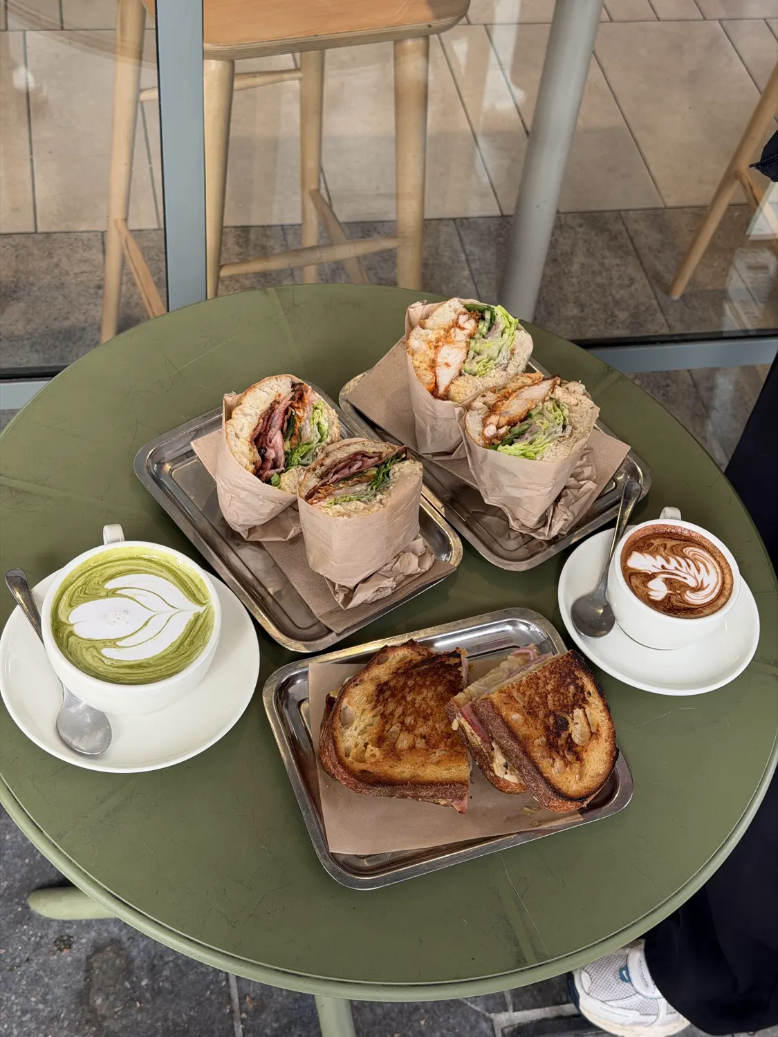 An overhead view of a shared table at Keens Sandwich Shop in Rosebery: two sandwiches on a tray, a toasted sandwich, a matcha latte and a cappuccino